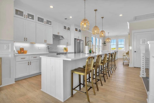 A modern kitchen with white cabinetry, a central island with bar stools, pendant lighting, and a large window. - NEW 5BR Suite | Steps to Beach w/Parking/Elevator! (Carolina Beach)