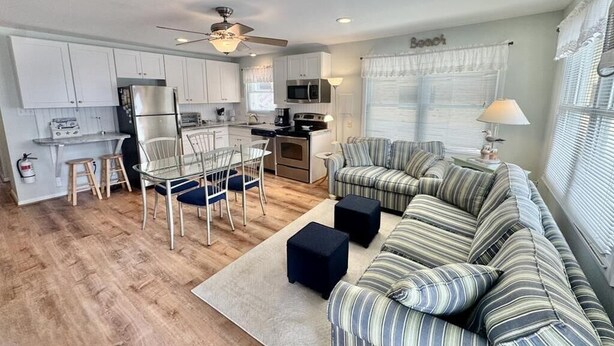 A modern kitchen with white cabinets, a dining area with a glass table and chairs, and a striped sofa in a living room. - Steps to the Sand: Bright, Spacious Condo on South-End Beach Block (Ocean City)