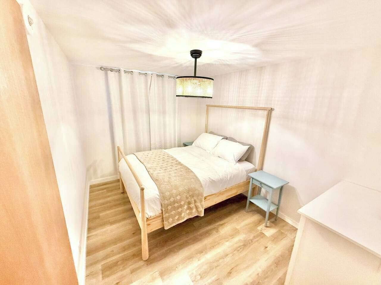 Un dormitorio con una cama de madera, una mesita de noche, una pequeña mesa, una lámpara colgante y una cortina blanca.