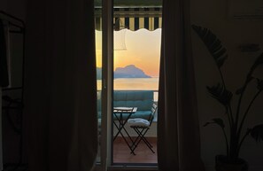 A view through a doorway to a balcony with a table and chairs, overlooking a body of water and a distant landmass during sunset. - Cephaluce Suite (Cefalù)