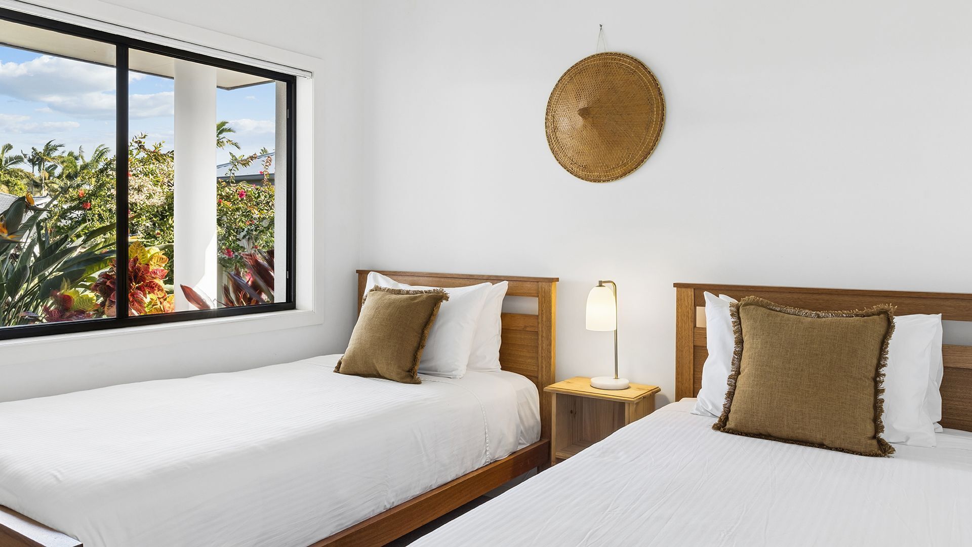 Une chambre d’hôtel avec deux lits, une fenêtre donnant sur une végétation tropicale, un panier tressé fixé au mur et une table de chevet avec une lampe.