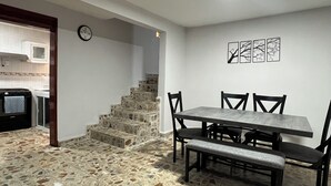 Un comedor con suelo de baldosas de piedra, una mesa con cuatro sillas, un banco y una escalera que sube a un nivel superior.