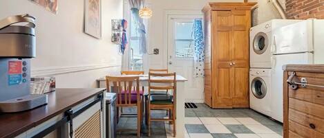 A compact kitchen with a coffee machine, a washing machine, and a dining area with chairs and a table.