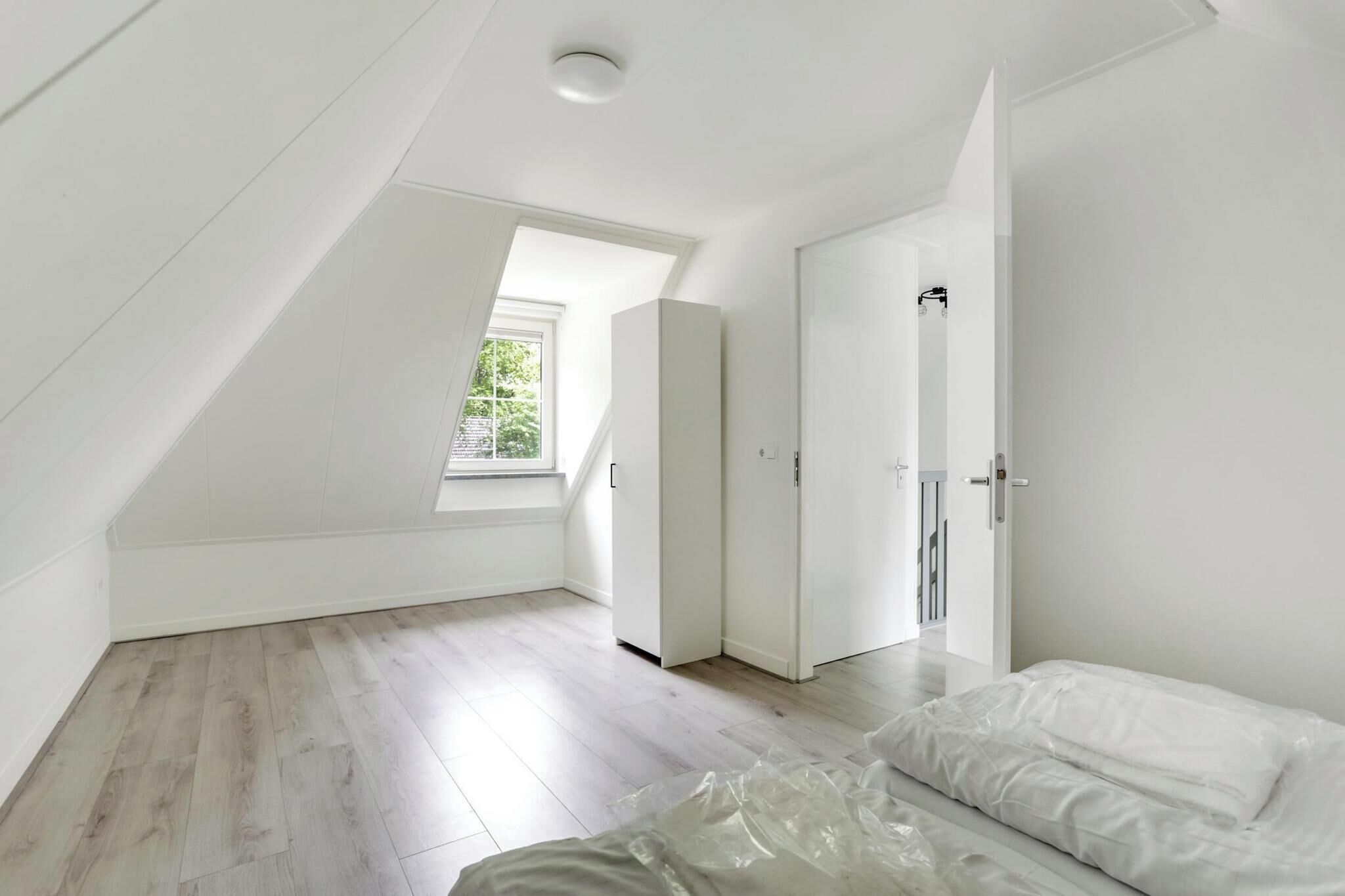 A room with a sloped ceiling, a window, a wardrobe, and a bed with a plastic cover.