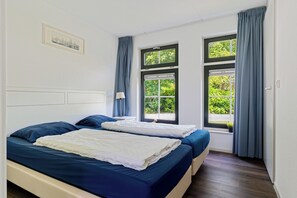 Ein Zimmer mit zwei Betten, einem Fenster mit Vorhängen, einem Nachttisch mit Lampe und einem gerahmten Bild an der Wand.