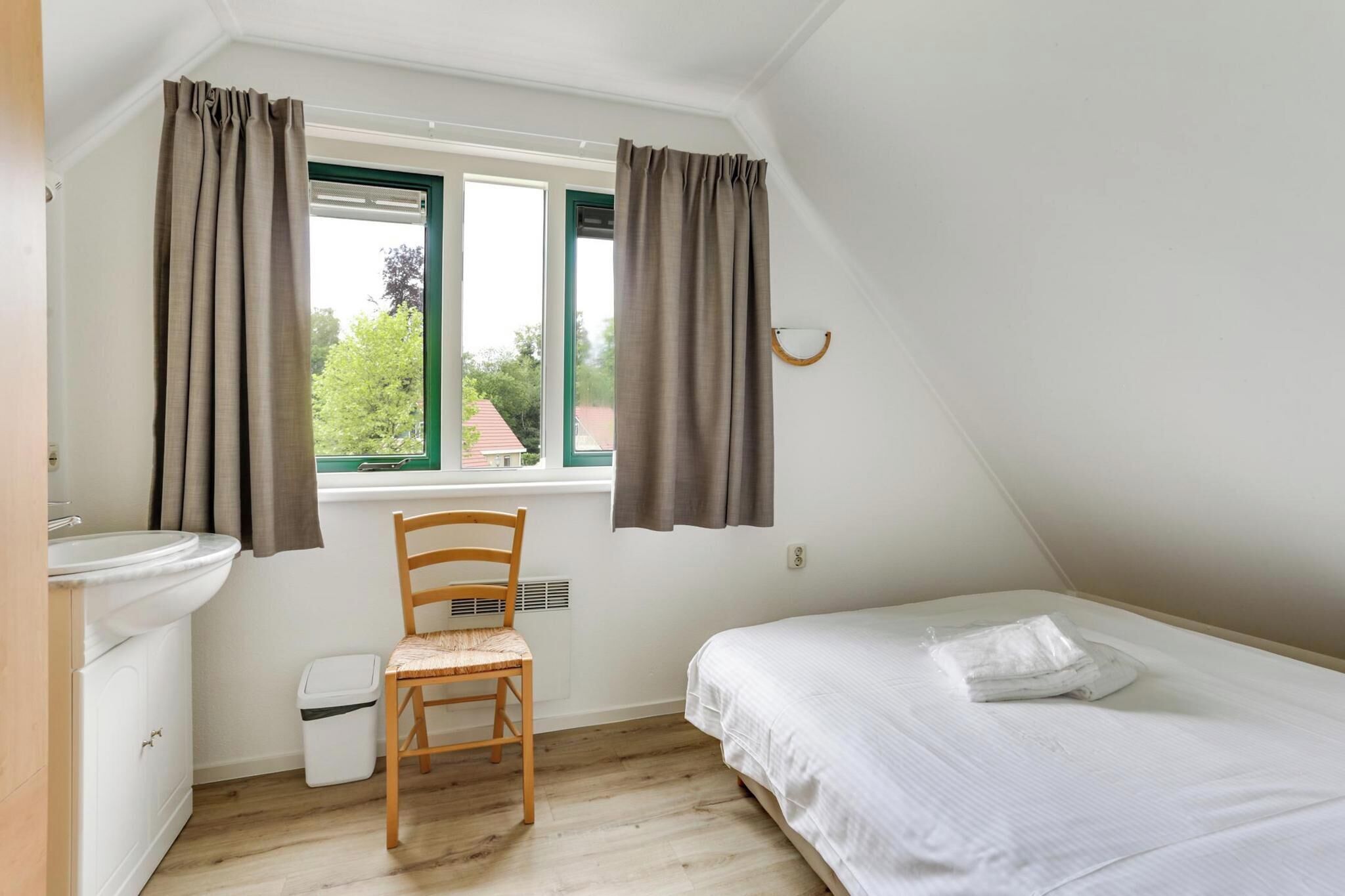 A small, clean room with a bed, a chair, a window with curtains, a sink, and a small trash bin.