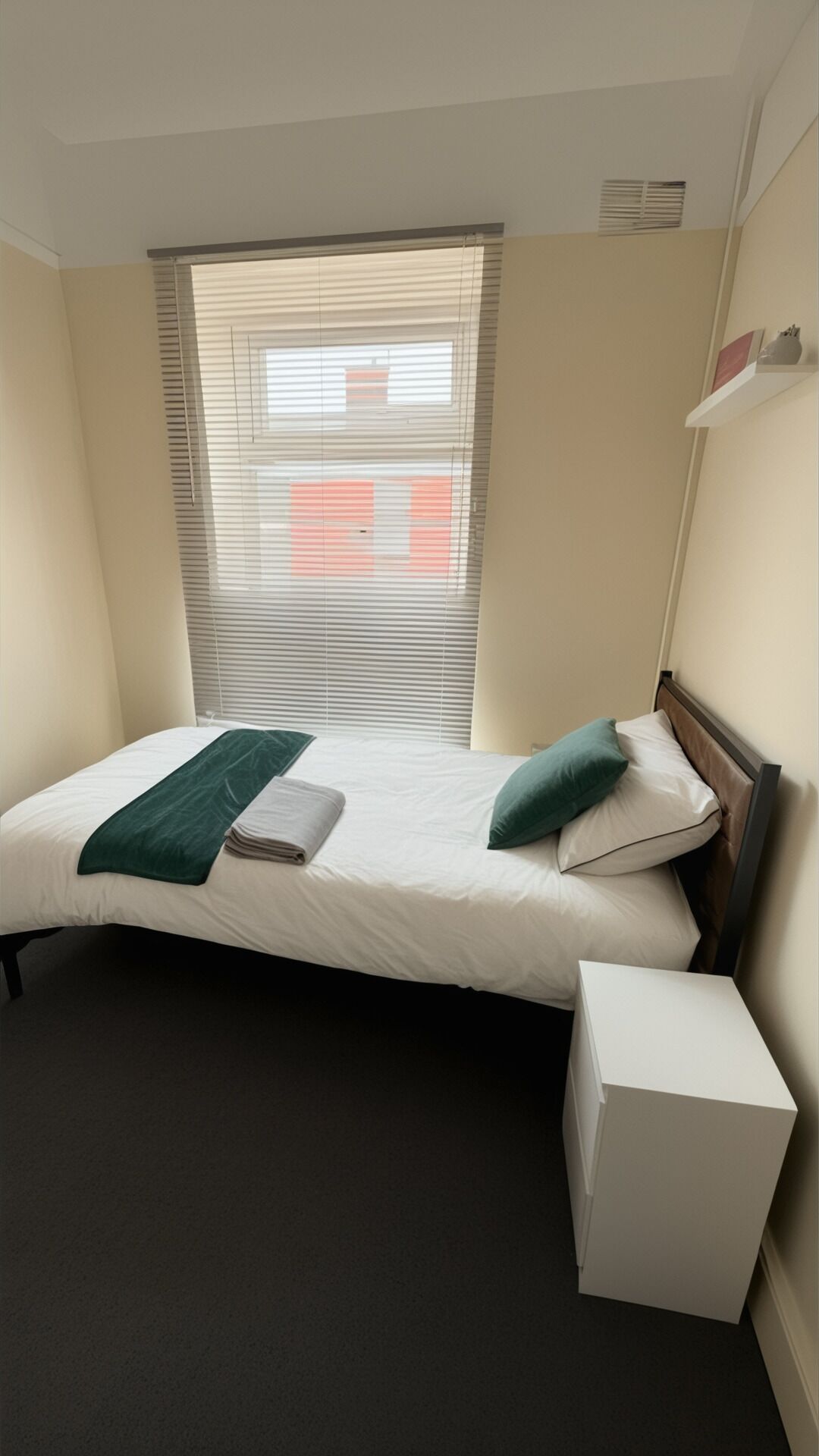 Un lit simple avec un couvre-lit vert, un oreiller et une table de chevet blanche, dans une pièce dotée d’une fenêtre et d’une grille d’aération.
