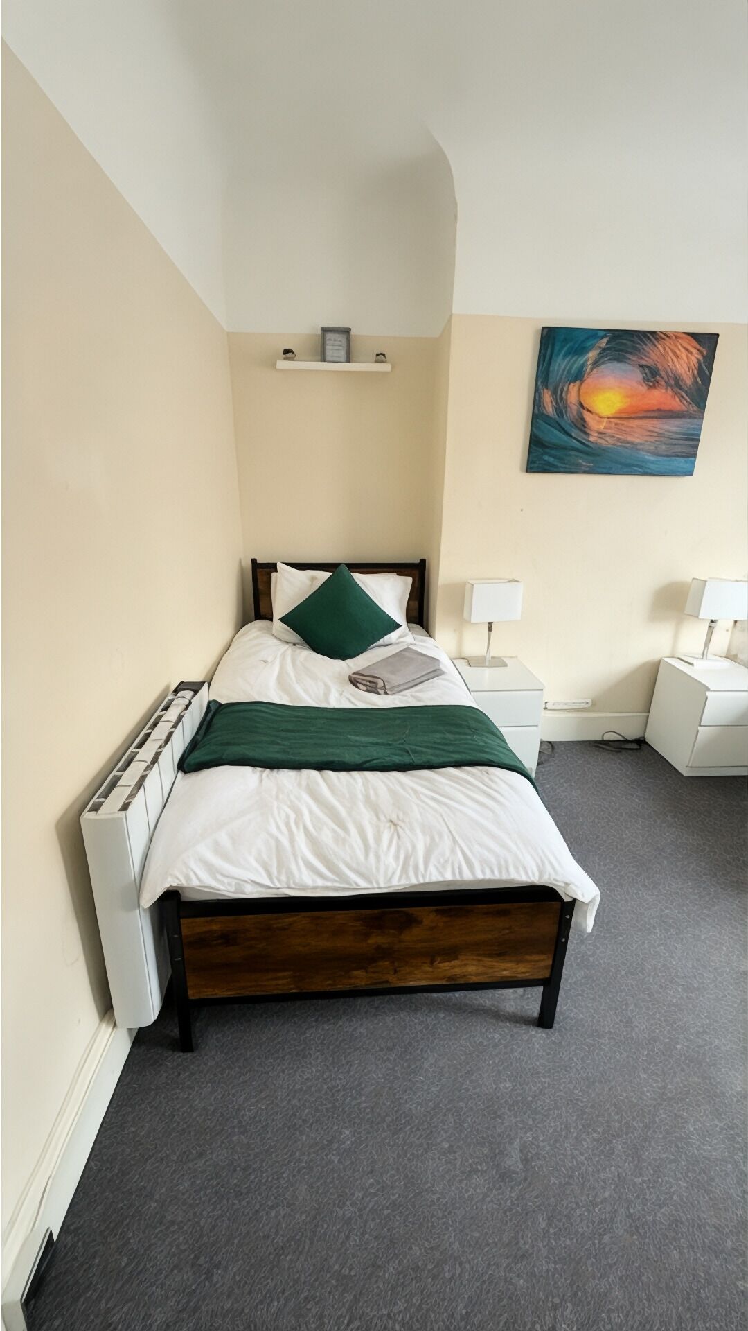 A neatly made bed with a green pillow, a bedside table with a lamp, a painting on the wall, and a small shelf above the bed.