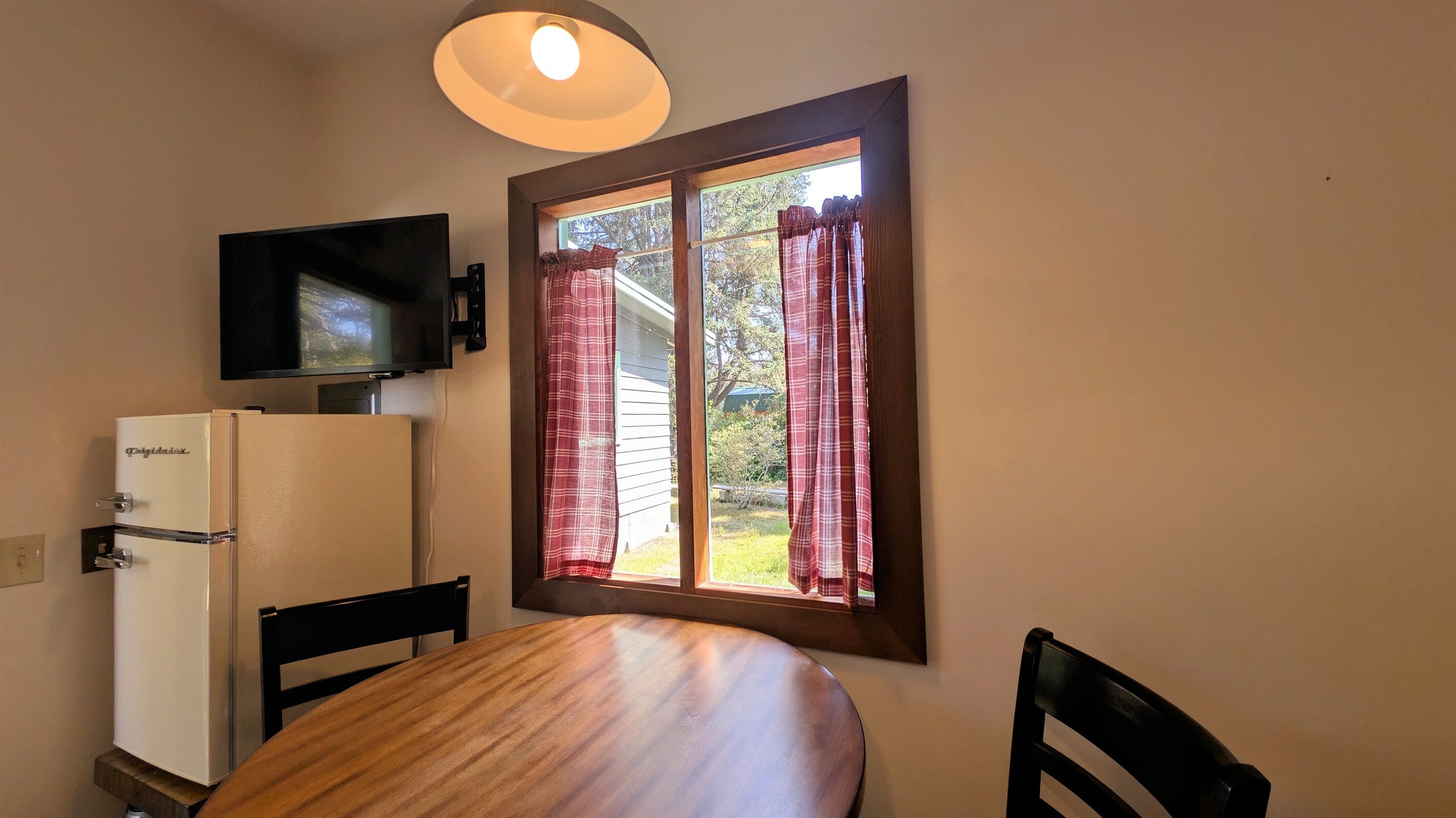 A compact room with a wooden table, a white refrigerator, a flat-screen TV mounted on the wall, and a window with plaid curtains.