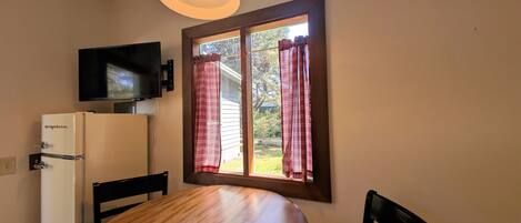 A compact room with a wooden table, a white refrigerator, a flat-screen TV mounted on the wall, and a window with plaid curtains.