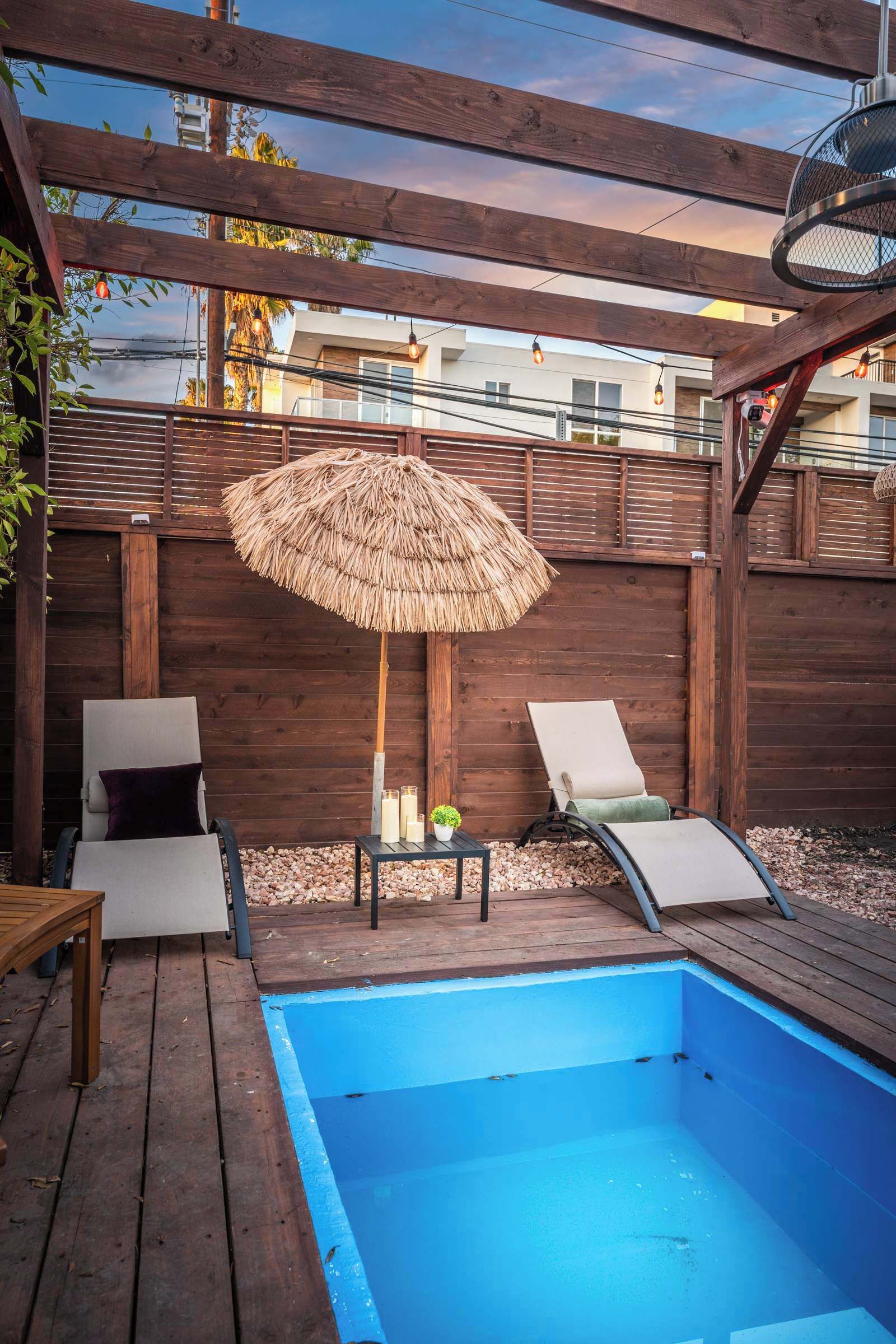 Una terraza de madera con una piscina azul, dos sillas de descanso, una mesa con velas y un paraguas de paja.