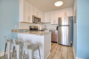 A kitchen with white cabinets, a stainless steel refrigerator, a microwave, and a built-in oven. There are two bar stools and a countertop.