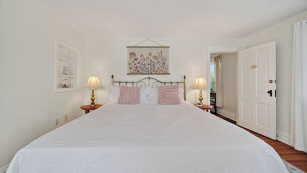 A bedroom with a large bed, two bedside tables with lamps, a wall-mounted artwork, and a door leading to another room.