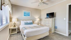 Un dormitorio con cama, dos mesitas de noche con lámparas, un aparador, un ventilador de techo, un televisor y un cuadro en la pared.