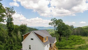 Una casa de estilo granero, de color blanco, con un porche de madera, rodeada de vegetación exuberante y montañas al fondo.