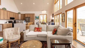 Amplia sala de estar con sofá, sillón y mesa de centro, cocina con armarios de madera y ventanas grandes con vistas.