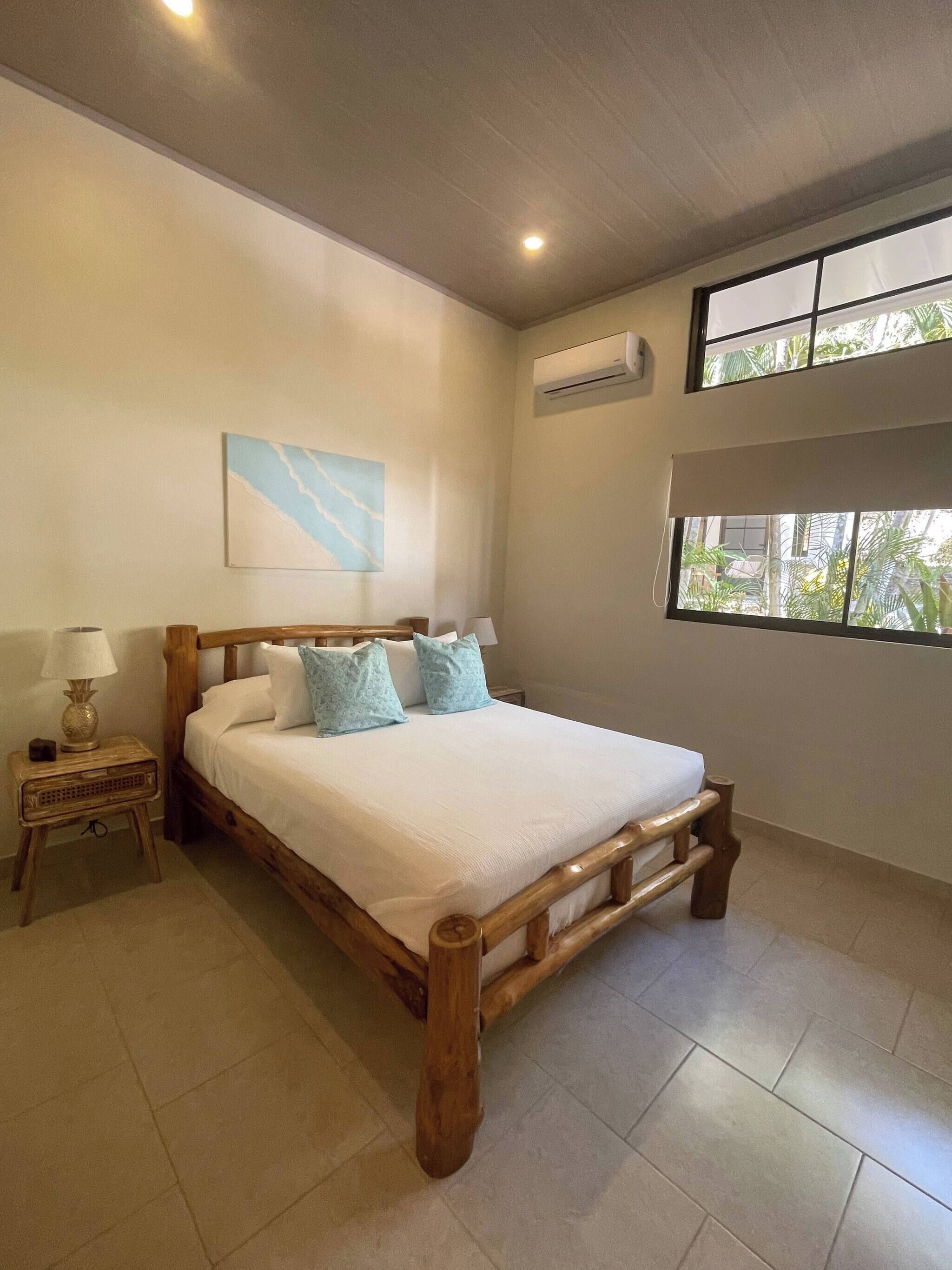 Un dormitorio con una cama de madera, una mesita de noche, aire acondicionado de pared y una ventana con vista a follaje.