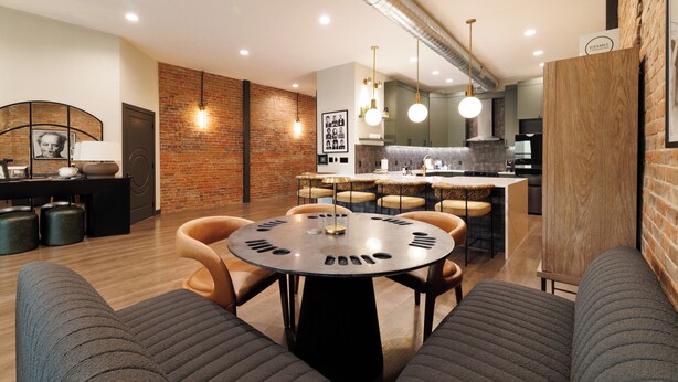 A modern interior with a round table, chairs, a kitchen area, and a brick wall. - The Aerie- On Central- LeMars (LeMars)