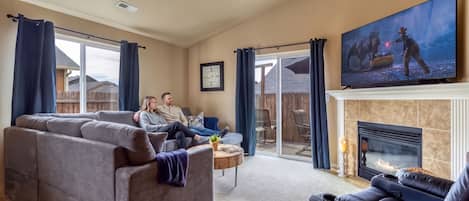 A living room with a large sectional sofa, a flat-screen TV, a fireplace, and a view through a sliding glass door.