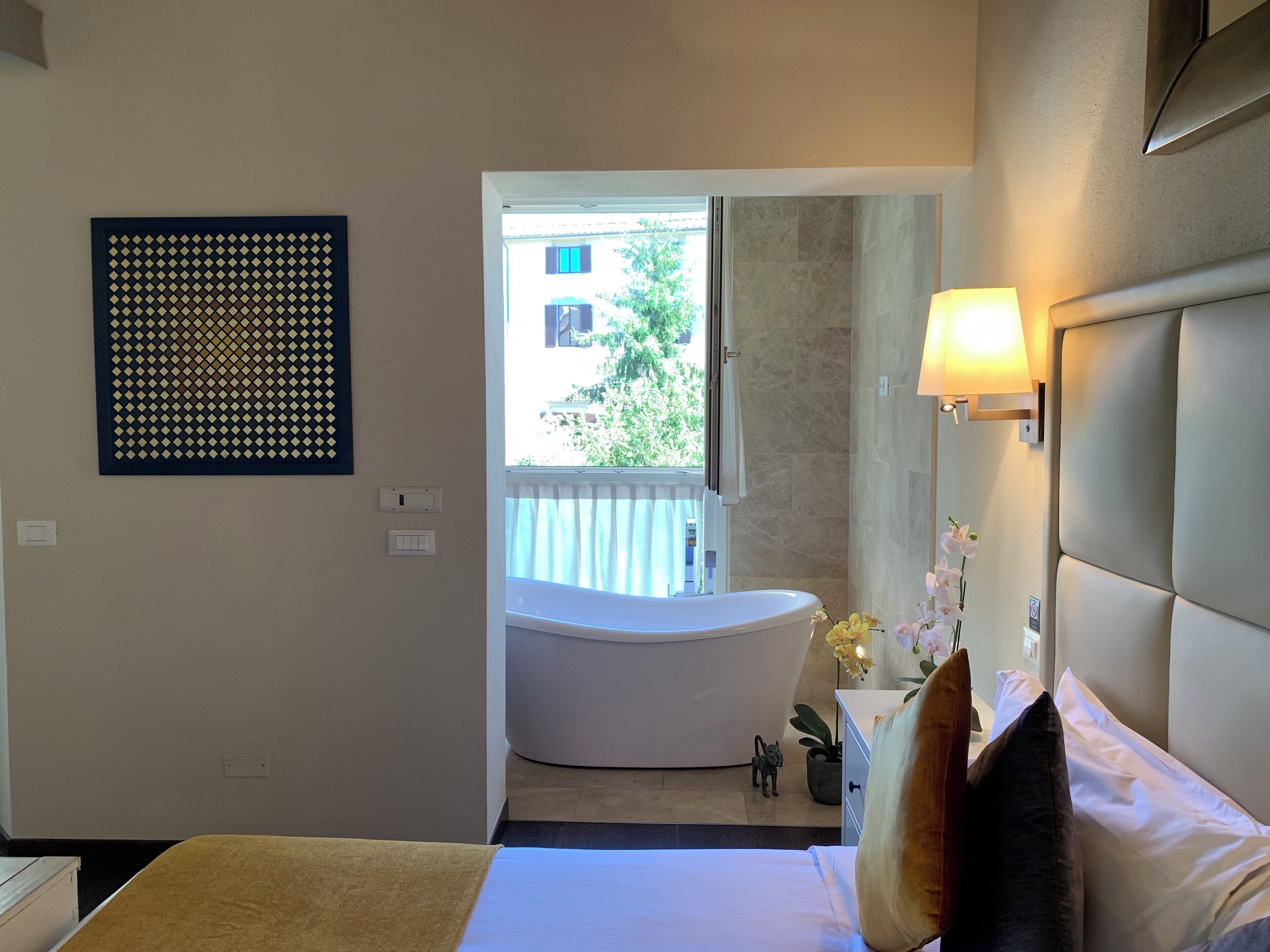 Una habitación de hotel con cama, baño con bañera, ventana con vistas a edificios y una lámpara fijada a la pared.