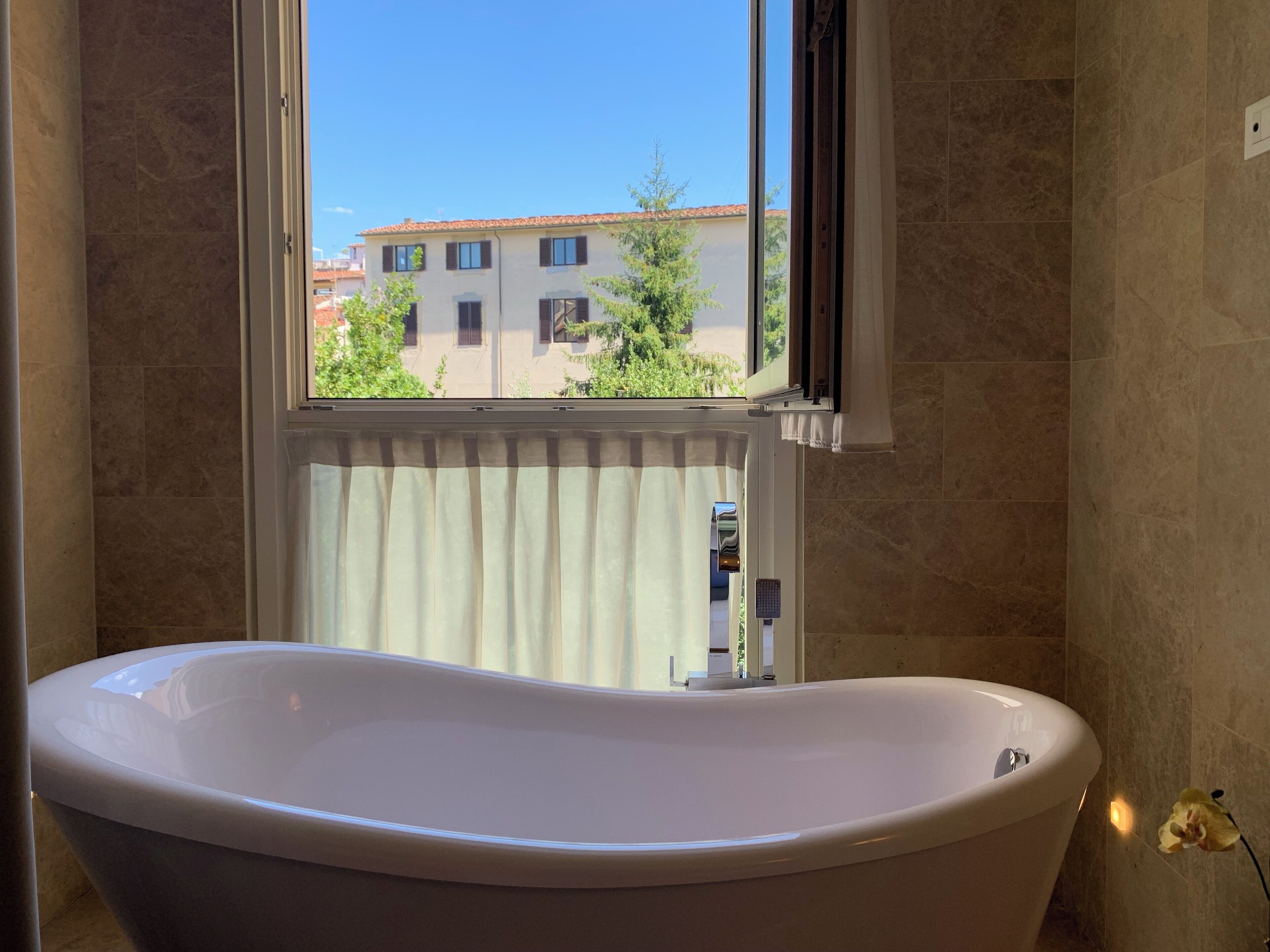 Ein Badezimmer mit freistehender Badewanne, einem Fenster mit Blick auf Gebäude und Bäume sowie einer Wandleuchte.