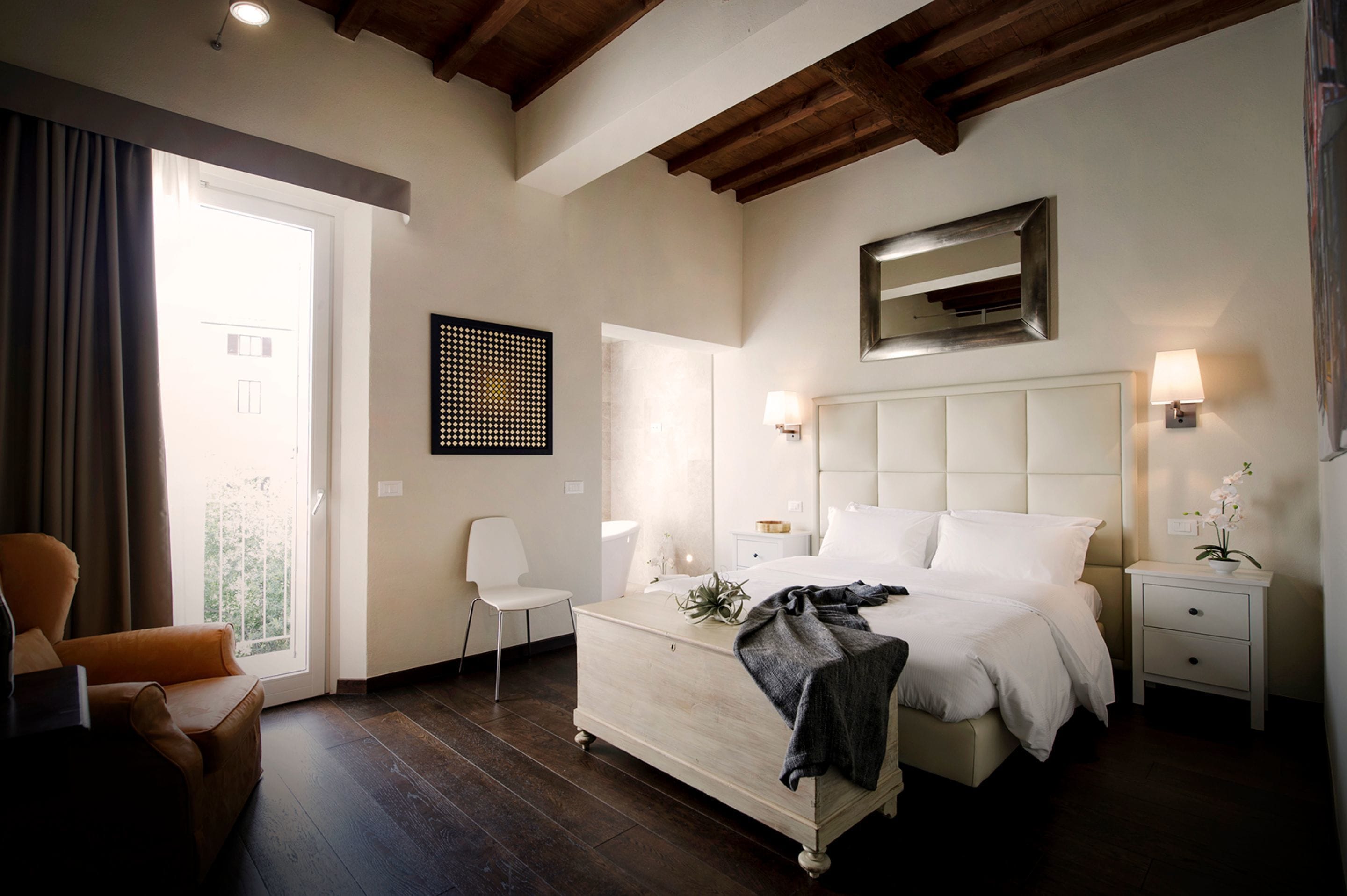 A bedroom with a large bed, a chair, a nightstand, a lamp, and a window with curtains.