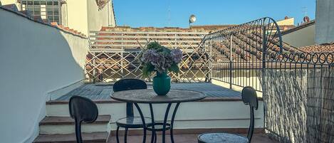 Una terraza en la azotea con una mesa, sillas, una planta en maceta y vistas a edificios al fondo.