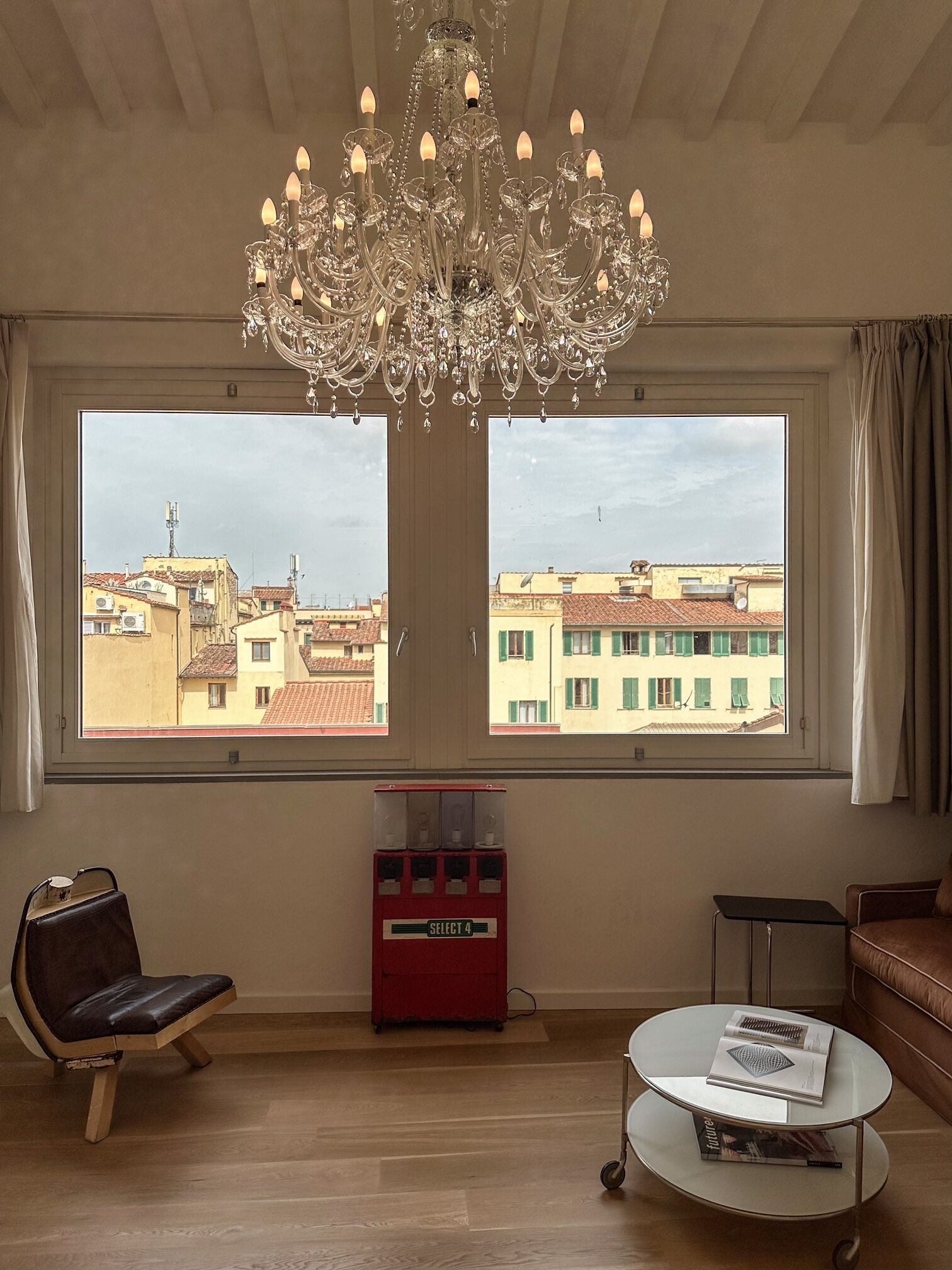 A room with a chandelier, a window with a view of buildings, a red vending machine, a leather chair, and a small table with magazines.