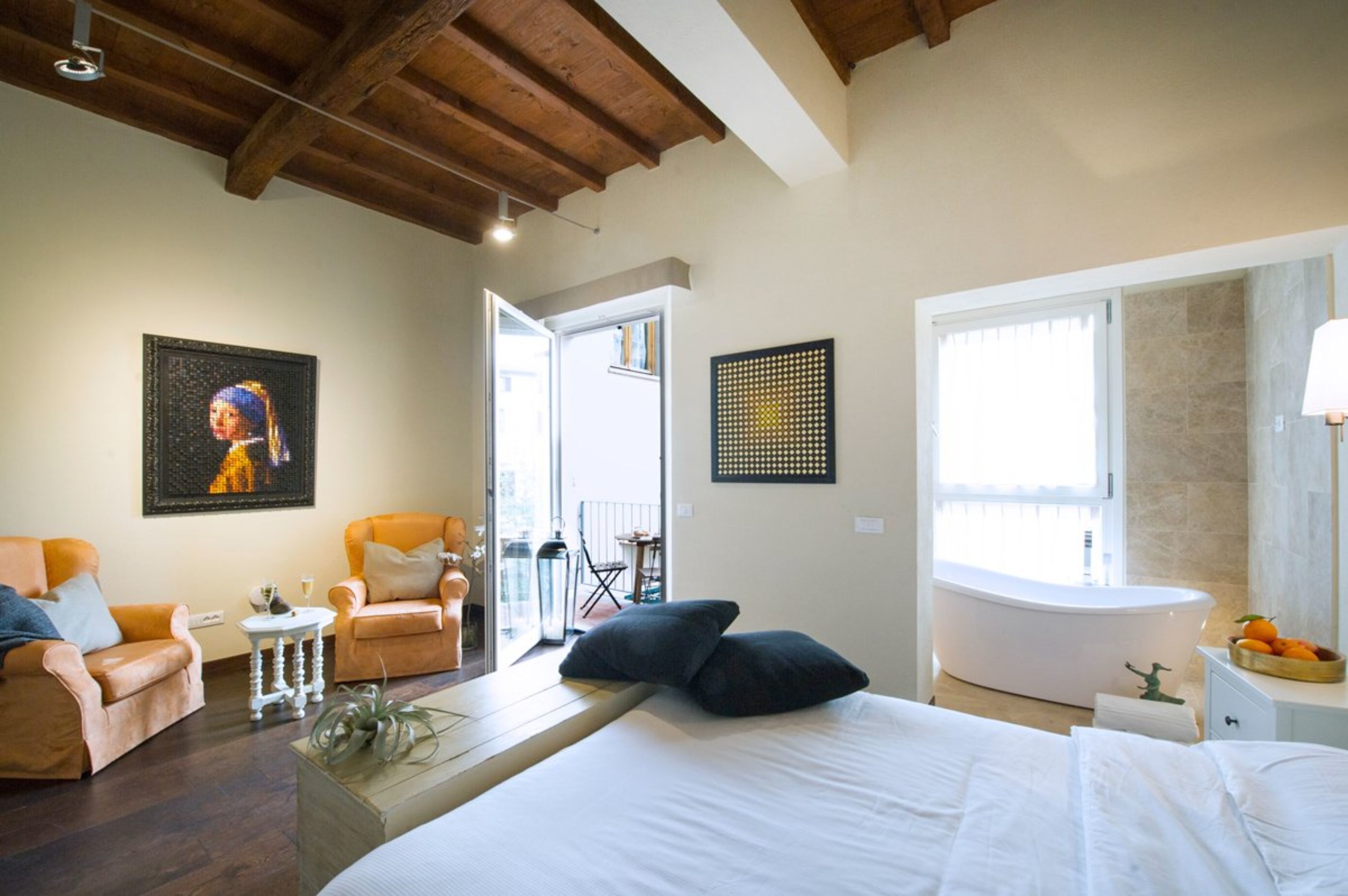 A bedroom with a large bed, two armchairs, a small table, a window with a view, a bathtub, and a door leading to a balcony.