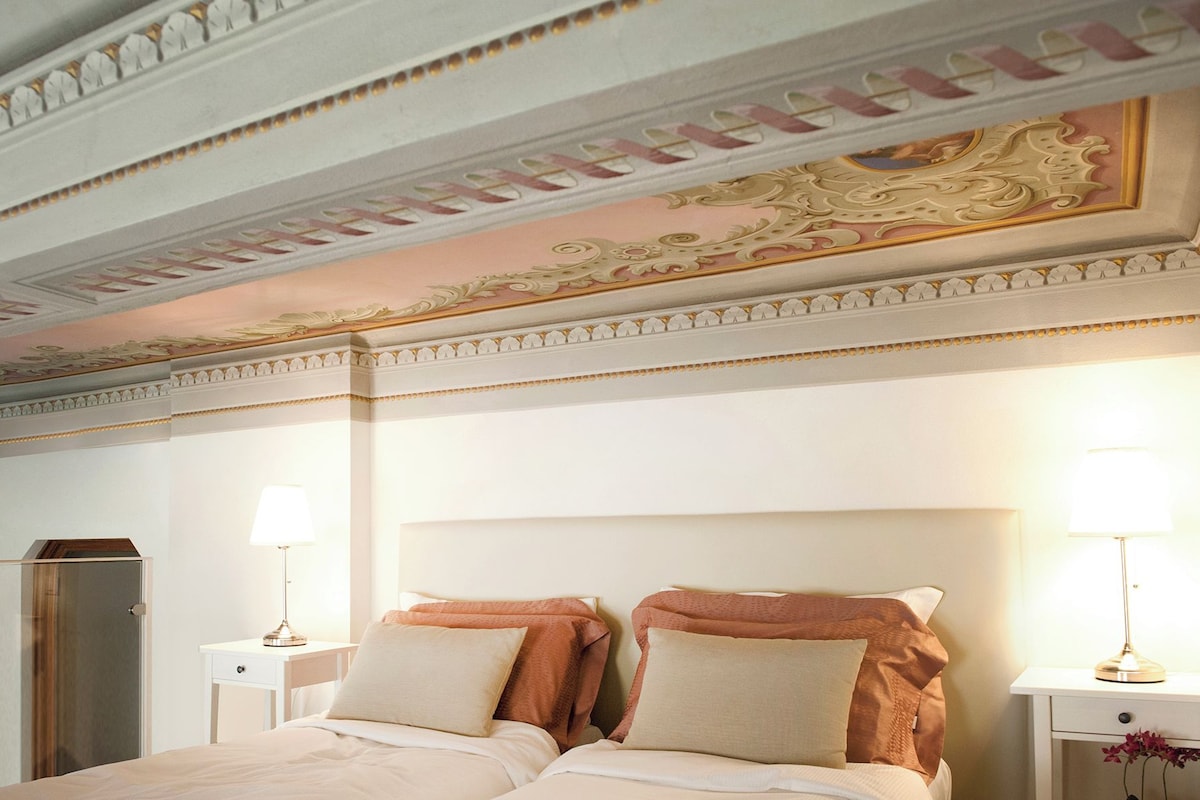 A hotel room with a large bed, two bedside tables with lamps, a wardrobe, and ornate ceiling decorations.