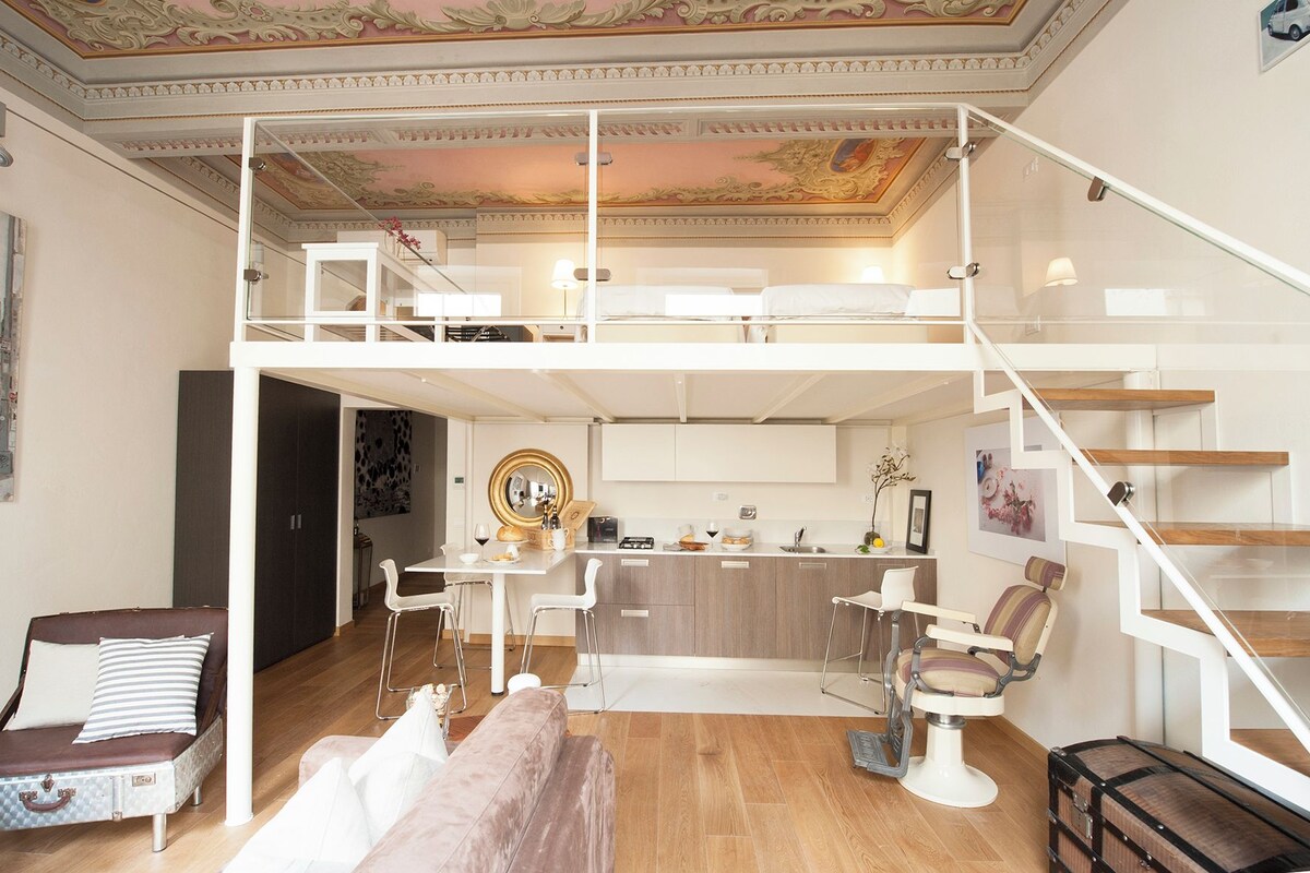 A spacious living area with a high ceiling, a dining table with chairs, a sofa, a small kitchen area, and a staircase.