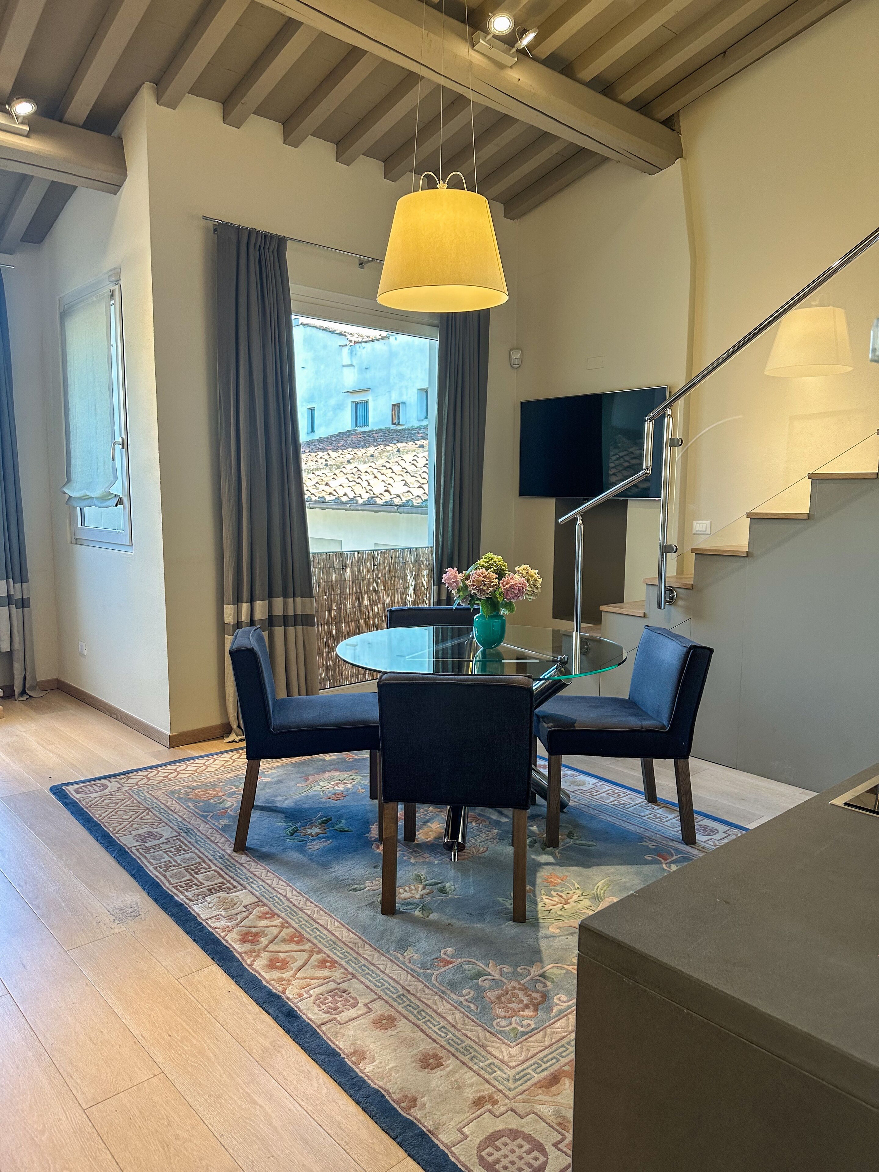 A modern living room with a glass dining table, dark chairs, a hanging lamp, a TV, and a staircase.
