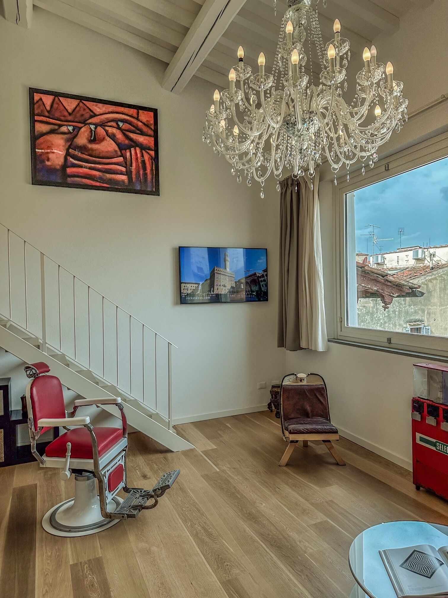 A room with a chandelier, a painting, a chair, a window with a view, and a red vending machine.