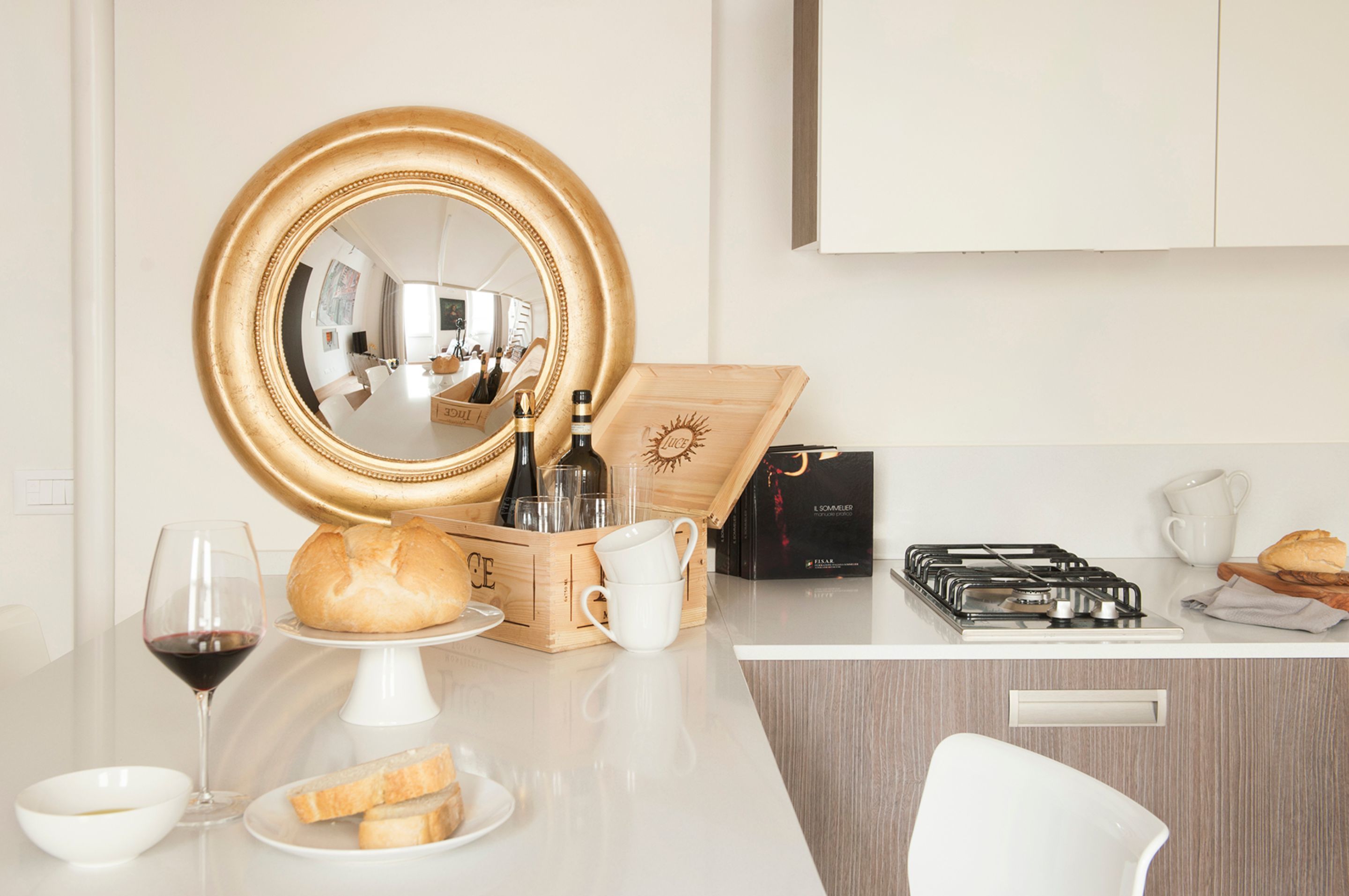 A kitchen counter with a mirror, a bread box, a wine glass, a loaf of bread, a white pitcher, a gas stove, and a white chair.