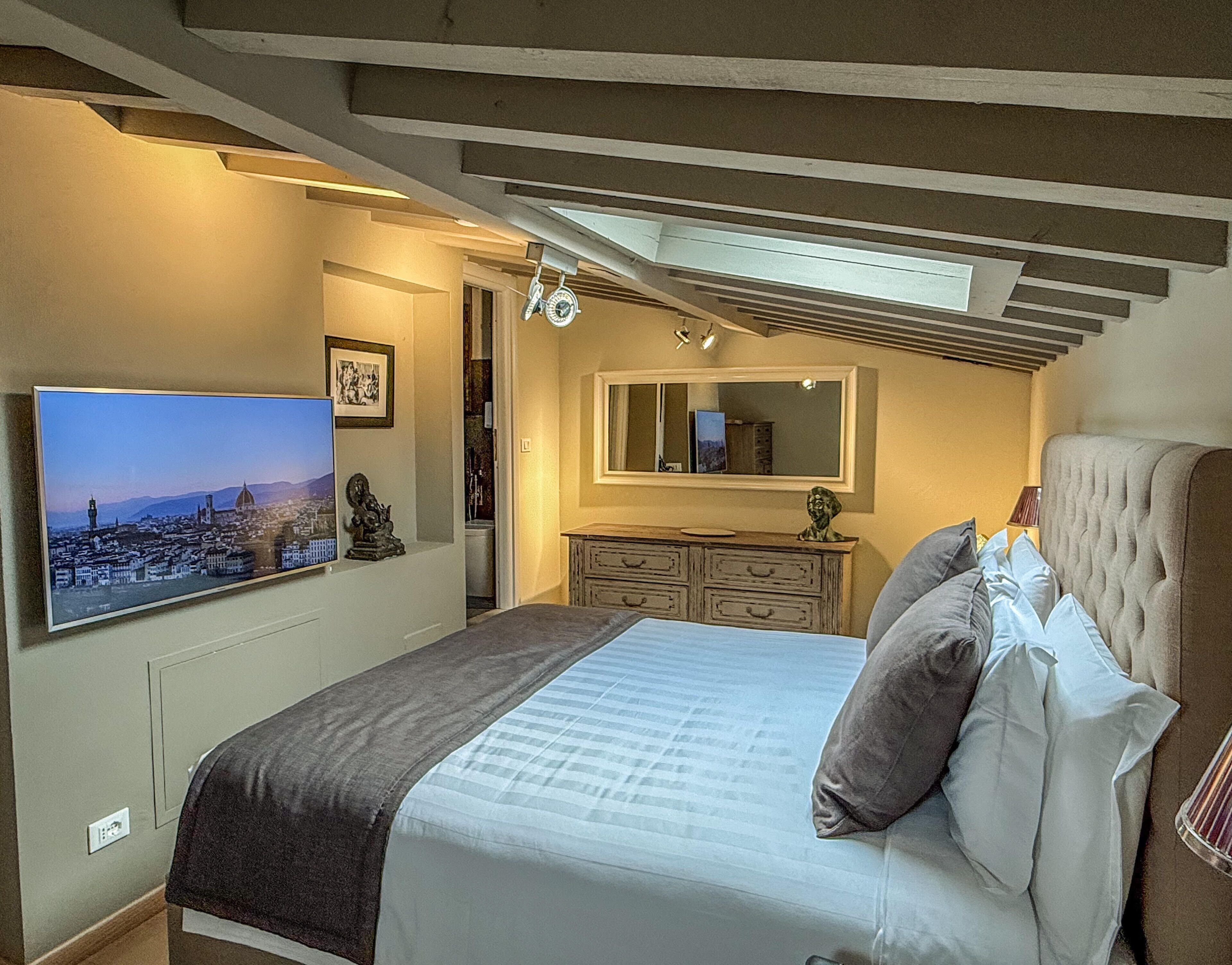Ein Schlafzimmer mit einem Bett, einem an der Wand montierten Fernseher, einem Schminktisch und einem Spiegel.
