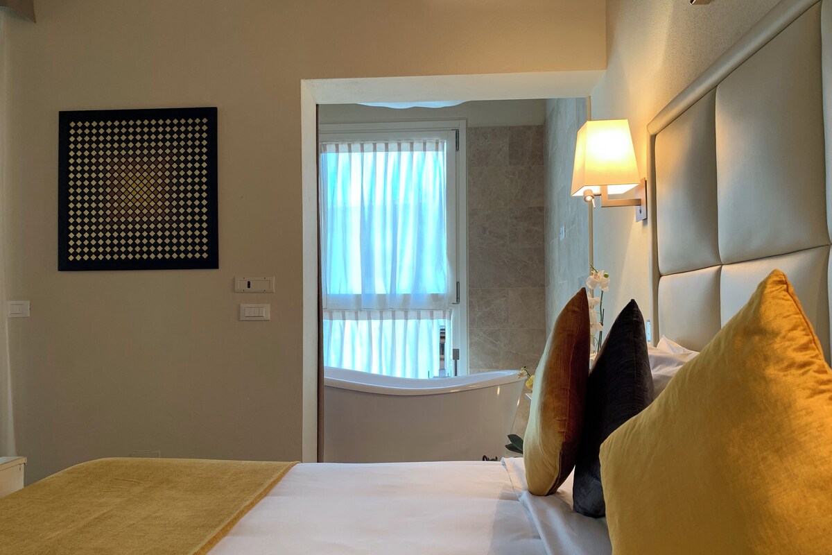 A hotel room with a bed, a window with curtains, a wall-mounted light, and a decorative panel.