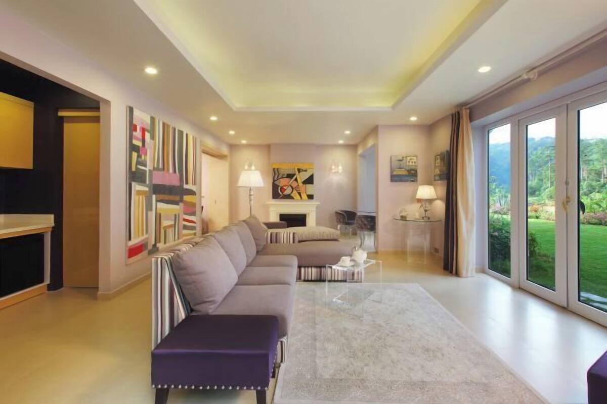 A modern living room with a sofa, a coffee table, and a fireplace. There is a large window providing a view of the outdoors.