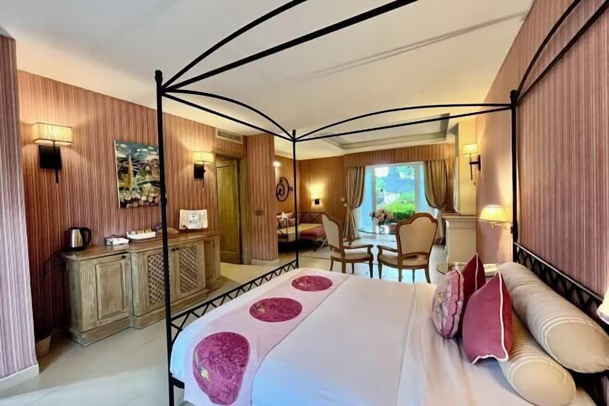 A hotel room with a four-poster bed, a wooden cabinet, a dining area with chairs, and a view of the outdoors.