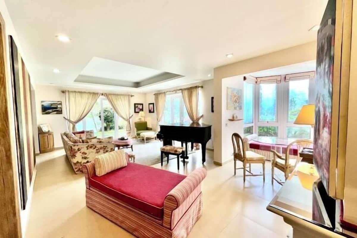A spacious room with a bed, a piano, a dining area, and large windows.