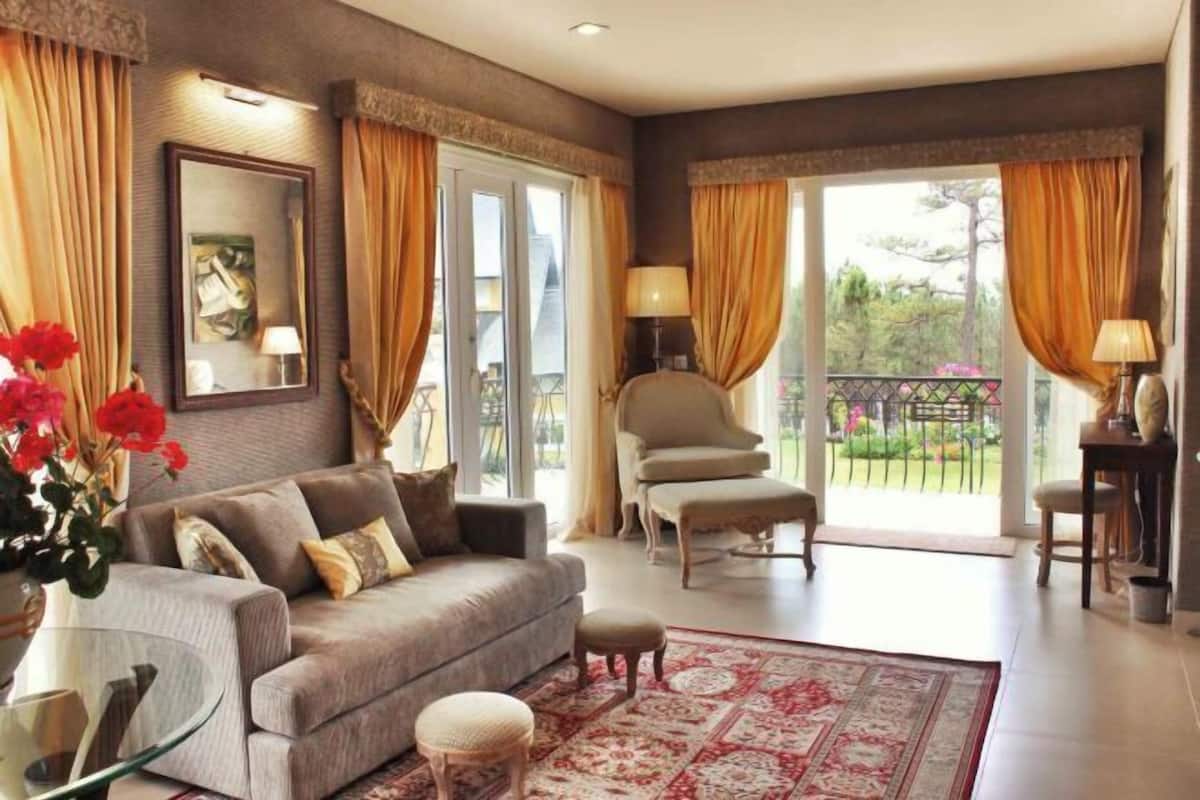 A living room with a sofa, ottoman, glass coffee table, and a patterned rug. There is a chair and a lamp near a window with curtains.