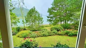 A view through a window of a garden with various plants and trees, leading to a body of water and hills in the distance. - Binh An Village Dalat Resort (Da Lat)