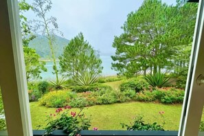 A view through a window of a garden with various plants and trees, leading to a body of water and hills in the distance. - Binh An Village Dalat Resort (Da Lat)