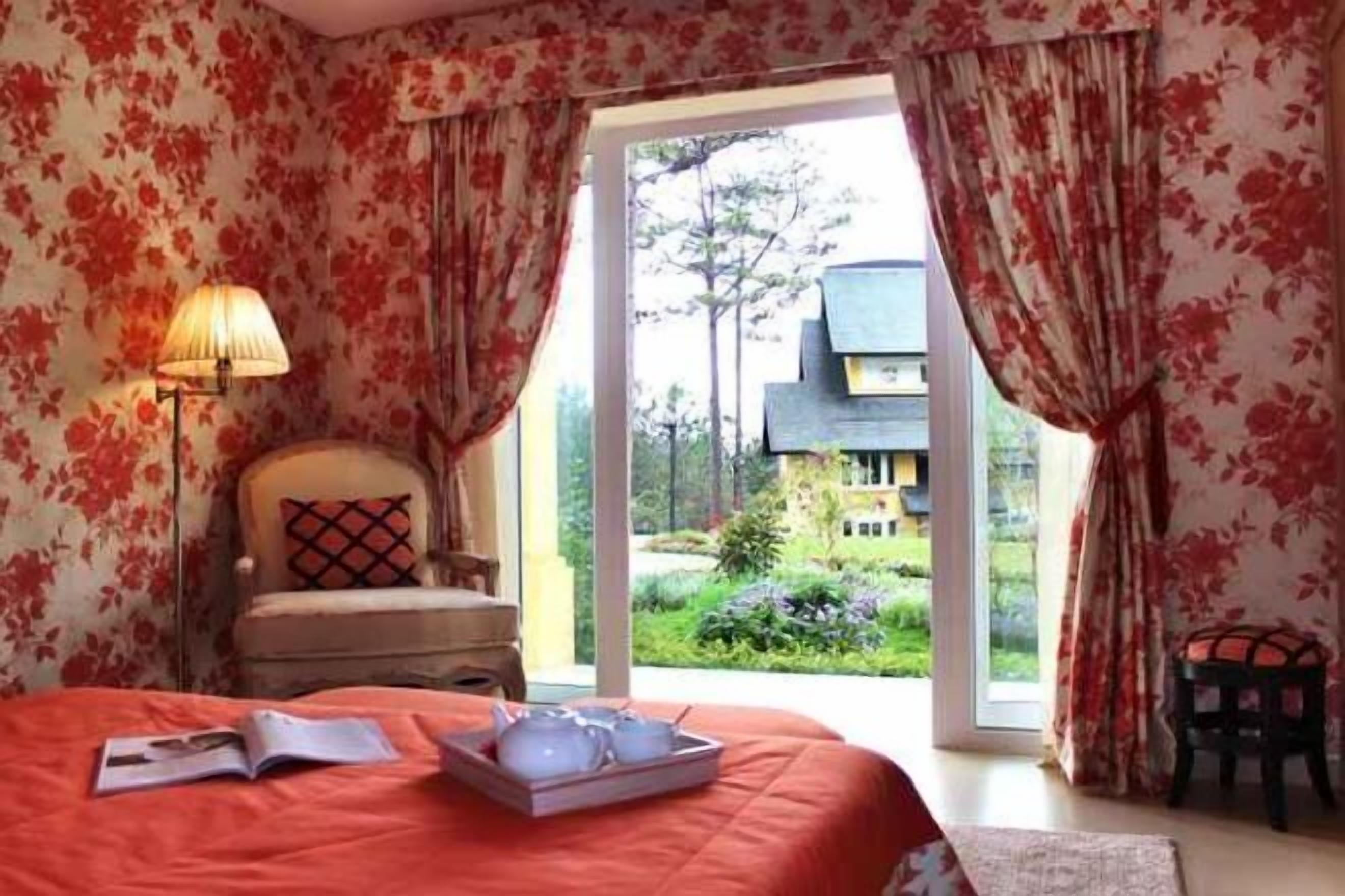 A bedroom with a bed, a chair, a table with a tray, and a view of a garden and building outside.