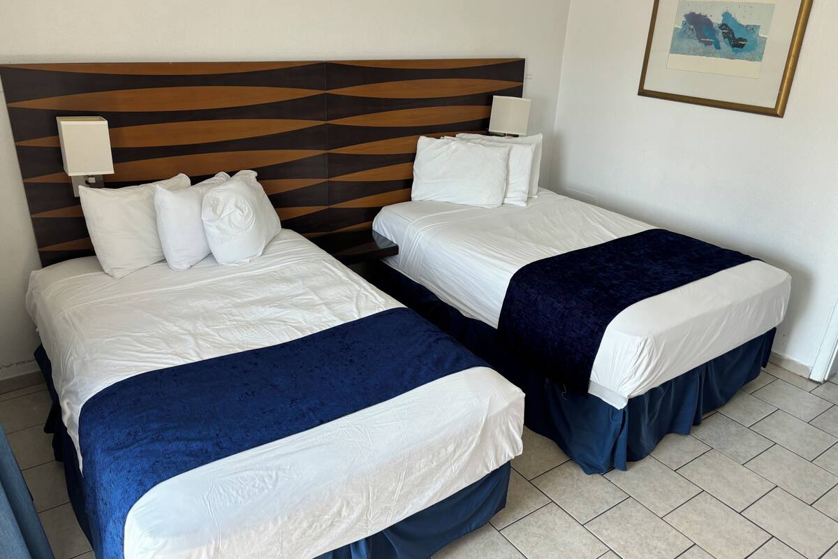 Habitación de hotel con dos camas, cada una con ropa blanca y detalles azules, un cabecero moderno de madera y una obra de arte enmarcada en la pared.