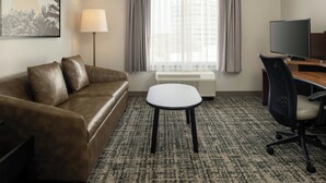 A hotel room with a brown leather sofa, a small white table, a black office chair, a flat-screen TV, a standing lamp, and a large window with sheer curtains. - Fairfield Inn & Suites Tampa Westshore / Airport (Tampa)