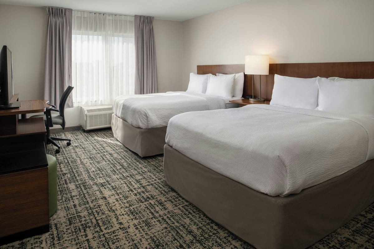 A hotel room with two beds, a desk, a chair, and a lamp.