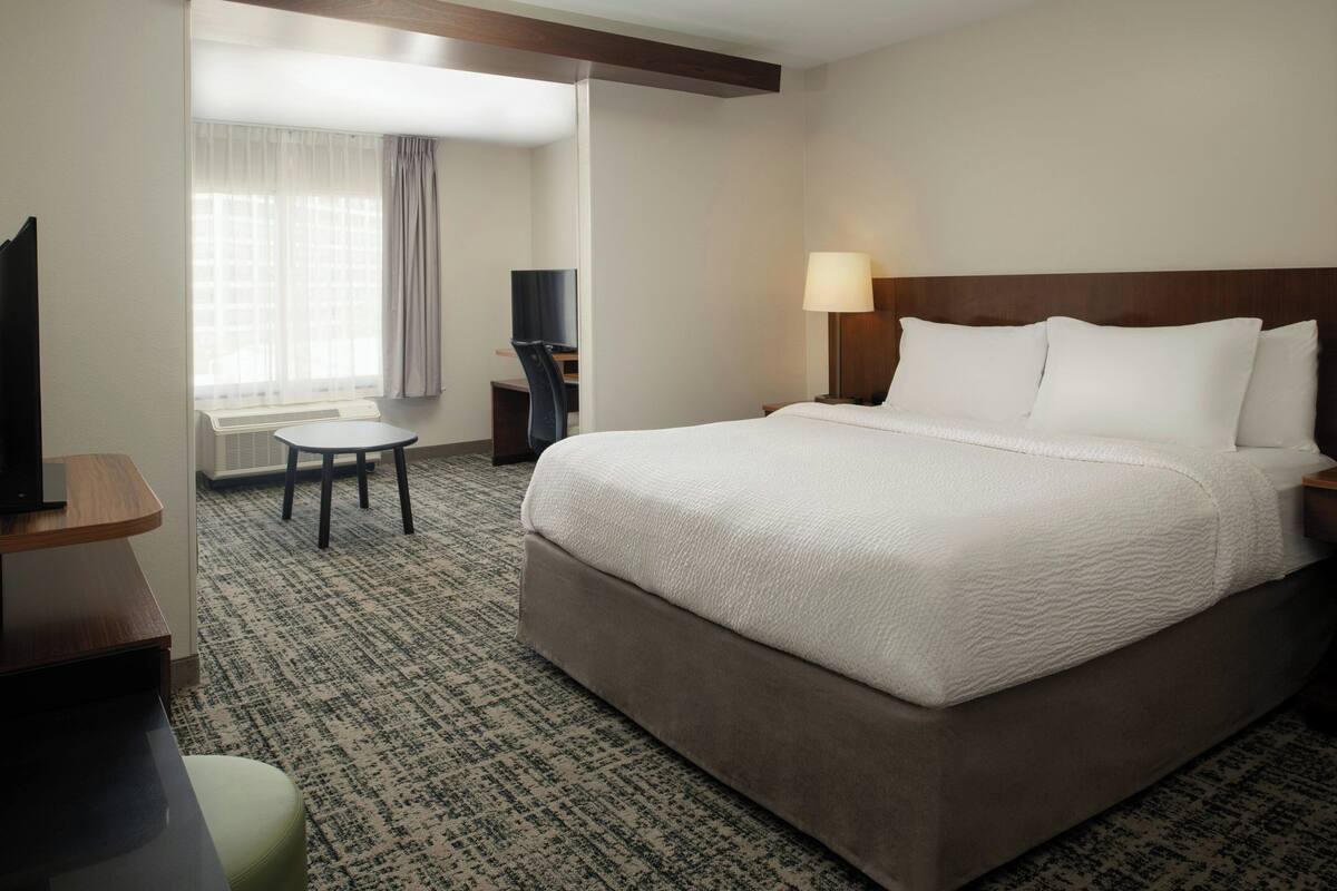 A hotel room with a large bed, a desk, a chair, a TV, and a lamp.