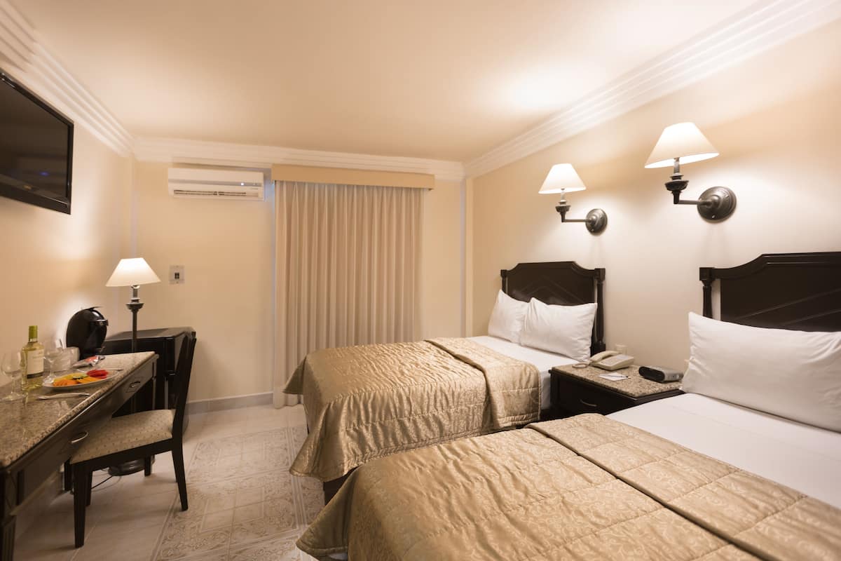 A hotel room with two beds, a desk with a lamp, a chair, and a television.