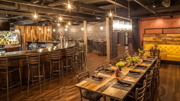 A restaurant with a long wooden table set for dining, bar area with stools, and a brick wall backdrop. - Warehouse Hotel (Manheim)