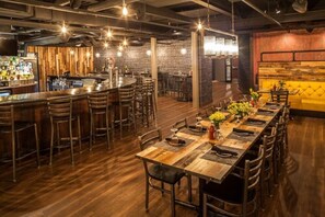 A restaurant with a long wooden table set for dining, bar area with stools, and a brick wall backdrop. - Warehouse Hotel (Manheim)